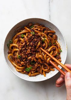 someone holding chopsticks over a bowl of noodles with ground beef and green onions