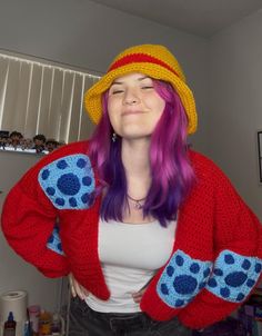 a woman with purple hair wearing a red cardigan and yellow hat is smiling at the camera