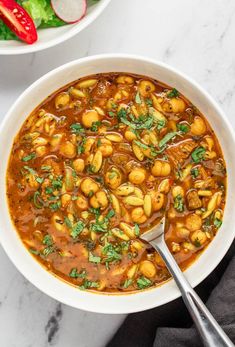 a white bowl filled with pasta and beans next to a salad on a marble table