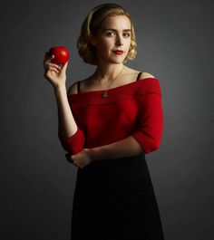 a woman holding an apple in her right hand and wearing a red sweater on top