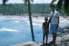 two people standing on rocks near the ocean with palm trees and beach in the background