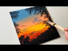 someone is holding a paintbrush and painting a sunset scene on a piece of canvas