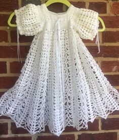 a white crochet baby dress hanging on a brick wall next to a yellow hanger