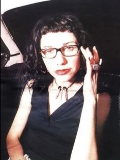 a woman wearing glasses sitting in a car with her hand up to her face and looking at the camera