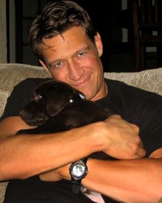 a man sitting on a couch holding a small dog in his arms and smiling at the camera