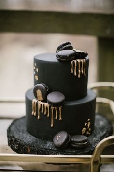 a three tiered black wedding cake with gold icing and macaroons on top