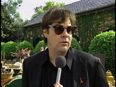 a man wearing sunglasses holding a microphone in front of a house with trees and bushes