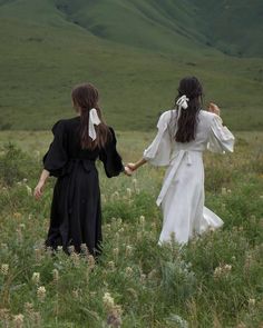 two girls in black and white dresses holding hands while walking through tall grass with mountains in the background