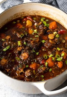 a pot filled with stew and vegetables on top of a blue table cloth next to a spoon