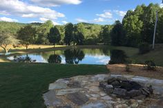 a stone fire pit in the middle of a grassy area next to a lake and trees