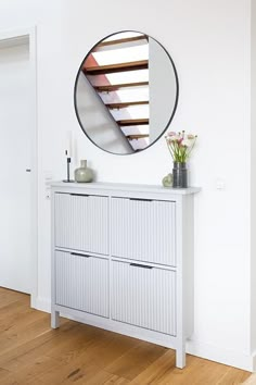 a white cabinet with drawers and a round mirror on the wall above it, in front of a wooden floor