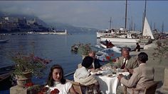 several people sitting at a table near the water with boats in the background and one person eating