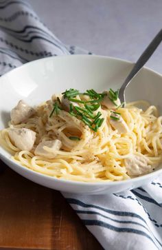 a white bowl filled with pasta and chicken on top of a wooden table next to a fork