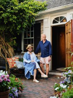 an older man and woman sitting on a bench in front of a house with flowers