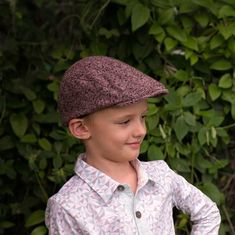 a young boy wearing a brown hat standing in front of some bushes and greenery