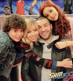 three people posing for the camera in front of a tv screen with other people behind them