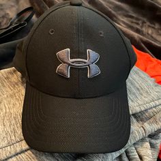 a gray and black under armour hat laying on top of a pile of clothing next to a pair of sunglasses