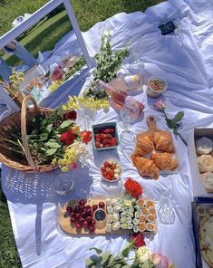 an outdoor picnic with food and drinks on the grass, including breads, croissants, fruit, and flowers