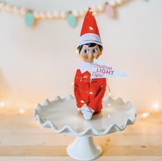 an elf is sitting on top of a cake plate and holding a light up sign