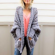 a woman standing in front of a white wall wearing a gray crochet cardigan