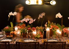 a table with candles, flowers and plates on it in front of a woman walking by