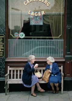 two older women sitting on a bench in front of a store window eating ice cream