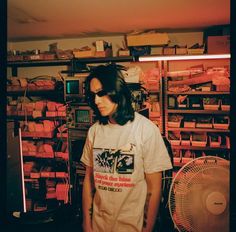 a man with long hair standing in front of a fan next to a shelf full of boxes