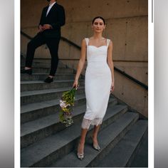 a woman in a white dress is walking down the stairs with flowers and a man standing behind her