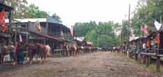 a group of people standing next to horses on a dirt road near buildings and trees