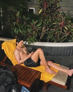 a shirtless man sitting on a yellow towel in front of a plant and wearing black shorts