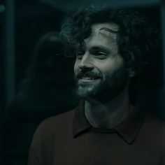 a man with curly hair and beard wearing a brown shirt smiles at the camera while standing in a dark room