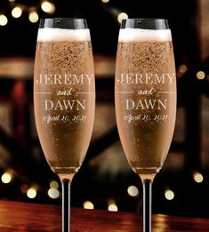 two champagne flutes sitting next to each other on top of a wooden table with lights in the background