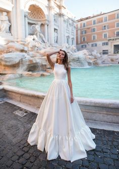 a woman in a white wedding dress standing next to a fountain and posing for the camera