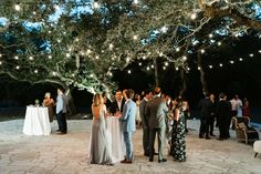 a group of people standing around each other at a dinner under a tree with lights