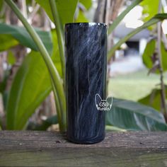 a black tumbler sitting on top of a wooden table next to green plants and trees