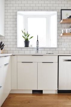 a white kitchen with wooden floors and open shelves on either side of the window, has a potted plant in it