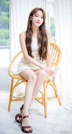 a young woman sitting on top of a wooden chair next to a white curtain and window