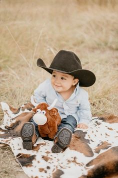 a little boy sitting on top of a cow wearing a cowboy hat and holding a stuffed animal