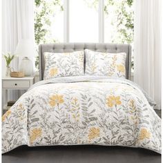 a bed with white and yellow comforters in front of a window next to a night stand