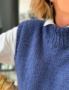 a woman wearing a blue sweater vest and gold hoop earrings with her hand on her hip