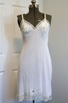 Gorgeous slip from the 1950s adorned with the most beautiful floral lace on the cups, hemline, and around the bust and top back. The hemline lace expands on the front to a larger triangular panel, adorned with the sweetest little embroidered white flowers with sage green leaves and stems. The bust cups have a three-flower lace panel with one embroidered flower in the centre. Adjustable straps with metal sliders. Falls approximately just below the knee (it's labelled "short", but keep in mind thi White Cami Dress With Lace Trim, Fitted Slip Dress With Lace Patchwork And Spaghetti Straps, Fitted Lace Patchwork Slip Dress For Weddings, White Lace Sleeveless Slip Dress, White Sleeveless Lace Slip Dress, White Slip Dress With Lace Trim And Spaghetti Straps, White Lace Slip Dress For Daywear, White Camisole Dress With Coquette Style, White Camisole Coquette Dress