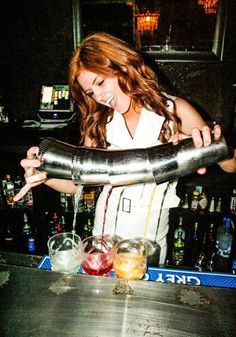 a woman is pouring drinks into a large metal pipe at a bar with liquor bottles in the background