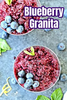 two glasses filled with blueberry granita on top of a gray surface next to green leaves