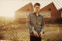 a young man standing in front of a fence with his hands in his pockets and looking at the camera