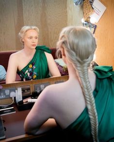 a woman in a green dress sitting at a desk with another person behind her looking into the mirror