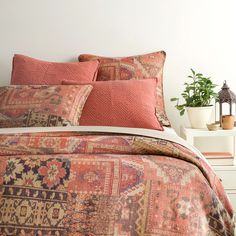 a bed covered in pink and orange pillows next to a white nightstand with potted plants