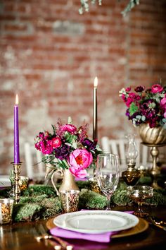 the table is set with purple flowers and gold plates, candles, and wine glasses