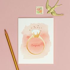 a pink card with a gold ring on it next to a pencil and stamp that reads congratulate