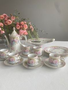 an assortment of porcelain dishes and cups on a table with pink roses in the background