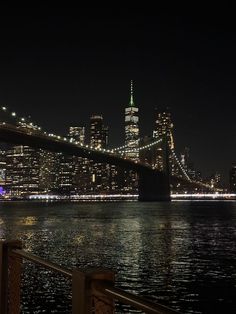 the city skyline is lit up at night as seen from across the water in front of the brooklyn bridge